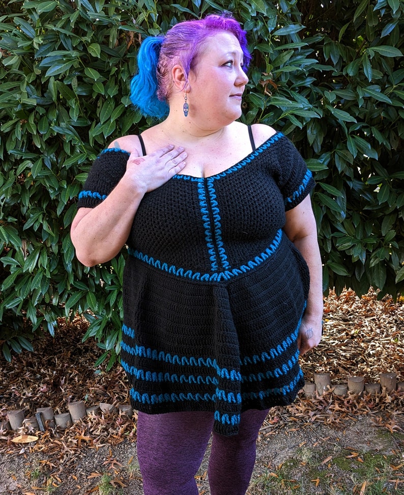 May include: Black crochet top with off-the-shoulder sleeves and a flared skirt. The top is accented with blue stitching along the neckline, sleeves, and skirt hem. The model is wearing purple leggings.