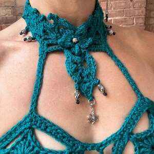 May include: A teal crochet halter top featuring a high neck collar with intricate floral patterns, silver-toned beads, and a dangling silver spiderweb charm. The garment is constructed with openwork stitching and decorative metal accents along the neckline.