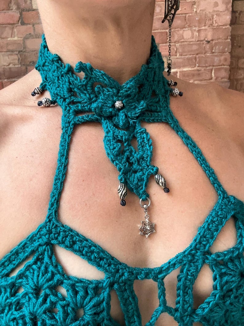 May include: A teal crochet halter top featuring a high neck collar with intricate floral patterns, silver-toned beads, and a dangling silver spiderweb charm. The garment is constructed with openwork stitching and decorative metal accents along the neckline.