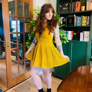 May include: A sleeveless, mustard yellow crochet dress with a flared skirt. The dress features a fitted bodice and a textured, open-weave design. The model is wearing black socks.