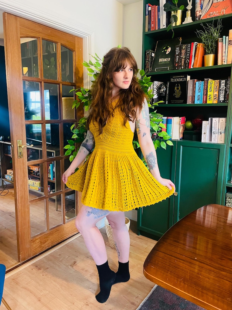 May include: A sleeveless, mustard yellow crochet dress with a flared skirt. The dress features a fitted bodice and a textured, open-weave design. The model is wearing black socks.