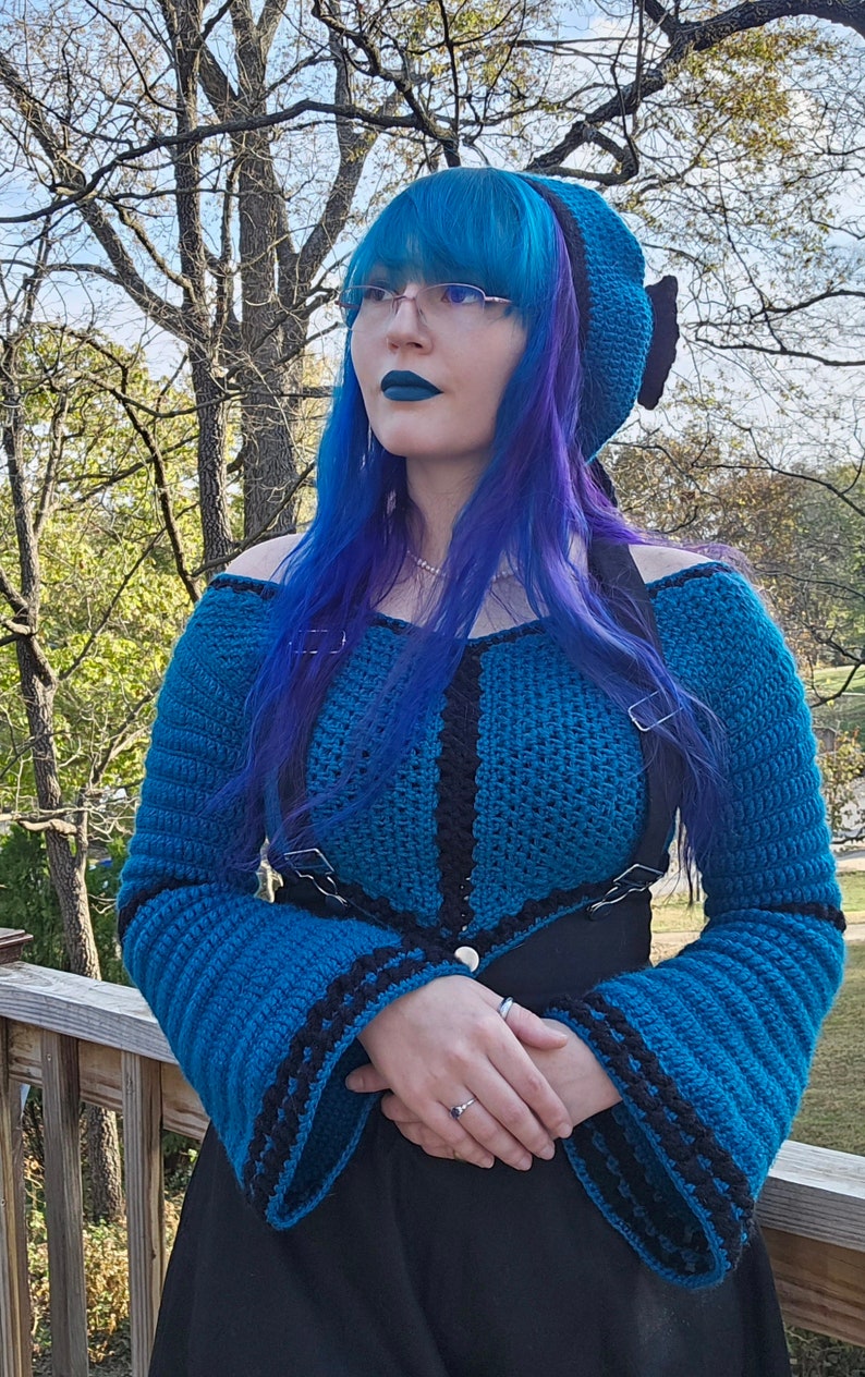 May include: A teal and black crochet top with long sleeves and a matching hat. The top features a unique design with black accents and a button closure. The hat has a black bow. The outfit is worn with a black skirt.