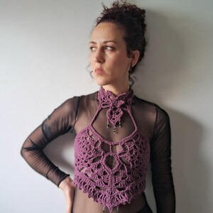 May include: A mauve crochet halter top featuring an intricate openwork pattern, a high neck collar, and small dangling bead accents. The garment is layered over a sheer black long-sleeve top, creating a textured look with a pointed hemline.