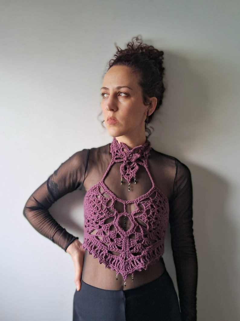 May include: A mauve crochet halter top featuring an intricate openwork pattern, a high neck collar, and small dangling bead accents. The garment is layered over a sheer black long-sleeve top, creating a textured look with a pointed hemline.