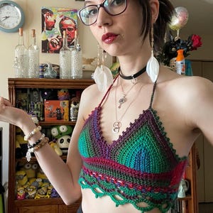 May include: A handmade crochet halter top in shades of green, purple, and red. The top features a scalloped edge and a halter neck tie. The top is worn with a black choker and multiple necklaces.