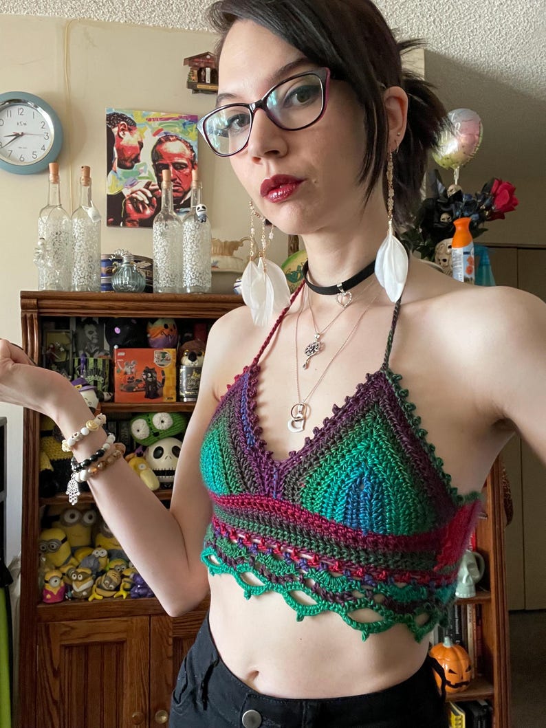 May include: A handmade crochet halter top in shades of green, purple, and red. The top features a scalloped edge and a halter neck tie. The top is worn with a black choker and multiple necklaces.