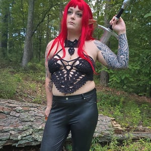 May include: A black crocheted halter top with an intricate geometric pattern, paired with black faux leather trousers. The outfit includes a long sword with a silver blade and a black hilt, worn over the shoulder, and bright red hair with pointed ears.