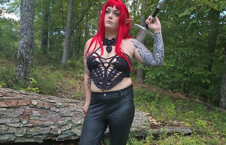 May include: A black crocheted halter top with an intricate geometric pattern, paired with black faux leather trousers. The outfit includes a long sword with a silver blade and a black hilt, worn over the shoulder, and bright red hair with pointed ears.