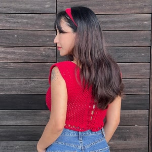 May include: A red crochet top with a matching headband and blue denim jeans. The sleeveless top has a detailed open-weave design and a lace-up back. The headband is a solid red color.