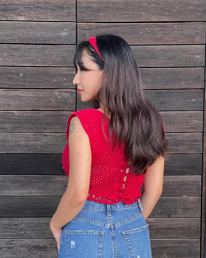 May include: A red crochet top with a matching headband and blue denim jeans. The sleeveless top has a detailed open-weave design and a lace-up back. The headband is a solid red color.
