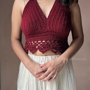 May include: A burgundy crochet halter top with a deep V-neck and scalloped hem. The top is handmade and has a textured appearance. The model is wearing a cream-colored skirt. The image includes the text "@metta threads".