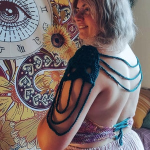 May include: A person wearing a green crocheted halter top with a matching crocheted shoulder piece. The top is tied at the back and has a layered design. The person is standing in front of a tapestry with a large eye design.