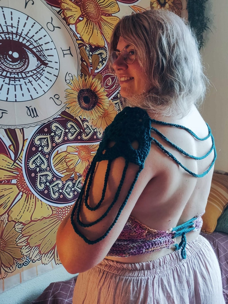 May include: A person wearing a green crocheted halter top with a matching crocheted shoulder piece. The top is tied at the back and has a layered design. The person is standing in front of a tapestry with a large eye design.