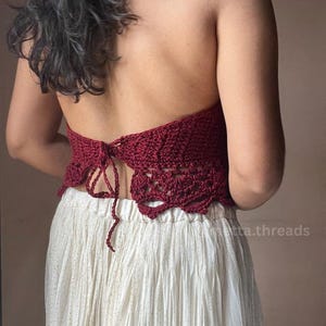 May include: A burgundy crochet halter top with a detailed openwork design. The top is tied at the back and paired with a cream-colored skirt. The skirt has a subtle gold shimmer.