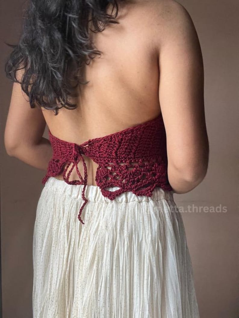 May include: A burgundy crochet halter top with a detailed openwork design. The top is tied at the back and paired with a cream-colored skirt. The skirt has a subtle gold shimmer.