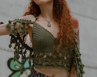 May include: A woman with long, curly red hair wearing a green crochet top with fringe detailing and a patterned skirt. She is wearing a silver necklace with a pendant and a colourful scrunchie on her wrist.