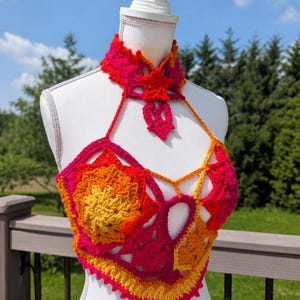 May include: A crochet halter top featuring a vibrant gradient of hot pink, orange, and yellow yarn. The garment includes a high-neck collar with a star-shaped accent, intricate floral-inspired motifs, and a pointed hemline with a scalloped edge.
