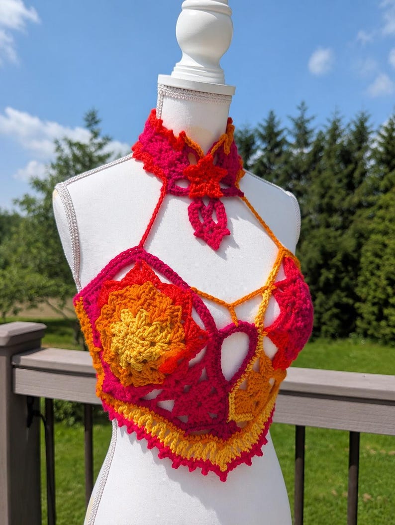 May include: A crochet halter top featuring a vibrant gradient of hot pink, orange, and yellow yarn. The garment includes a high-neck collar with a star-shaped accent, intricate floral-inspired motifs, and a pointed hemline with a scalloped edge.