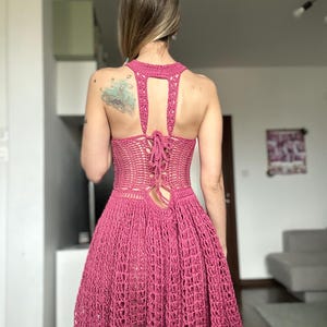 May include: A handmade, magenta-colored crochet dress. The dress features a halter neckline, open back with a lace-up closure, and a flared skirt. The crochet work has an open, airy design, perfect for warm weather. The dress is a vibrant, eye-catching piece.