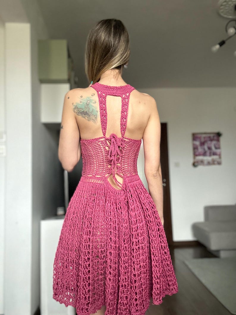 May include: A handmade, magenta-colored crochet dress. The dress features a halter neckline, open back with a lace-up closure, and a flared skirt. The crochet work has an open, airy design, perfect for warm weather. The dress is a vibrant, eye-catching piece.