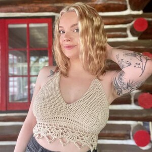 May include: A beige crochet halter top with a V-neck and scalloped hem. The top is handmade and has a textured appearance. The model is wearing the top with black denim shorts.