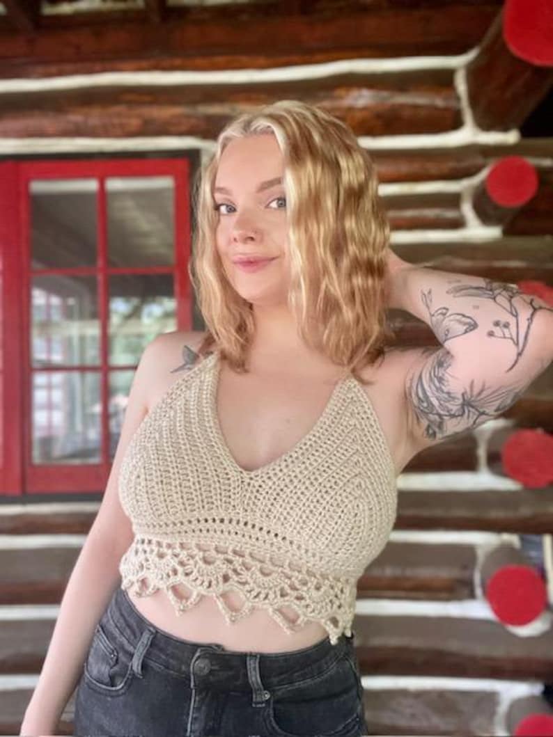 May include: A beige crochet halter top with a V-neck and scalloped hem. The top is handmade and has a textured appearance. The model is wearing the top with black denim shorts.