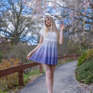 May include: A white and purple ombre crochet dress with short sleeves. The dress has a V-neck and a flared skirt. The model is wearing beige sandals. The background includes a path, a wooden fence, and flowering trees.