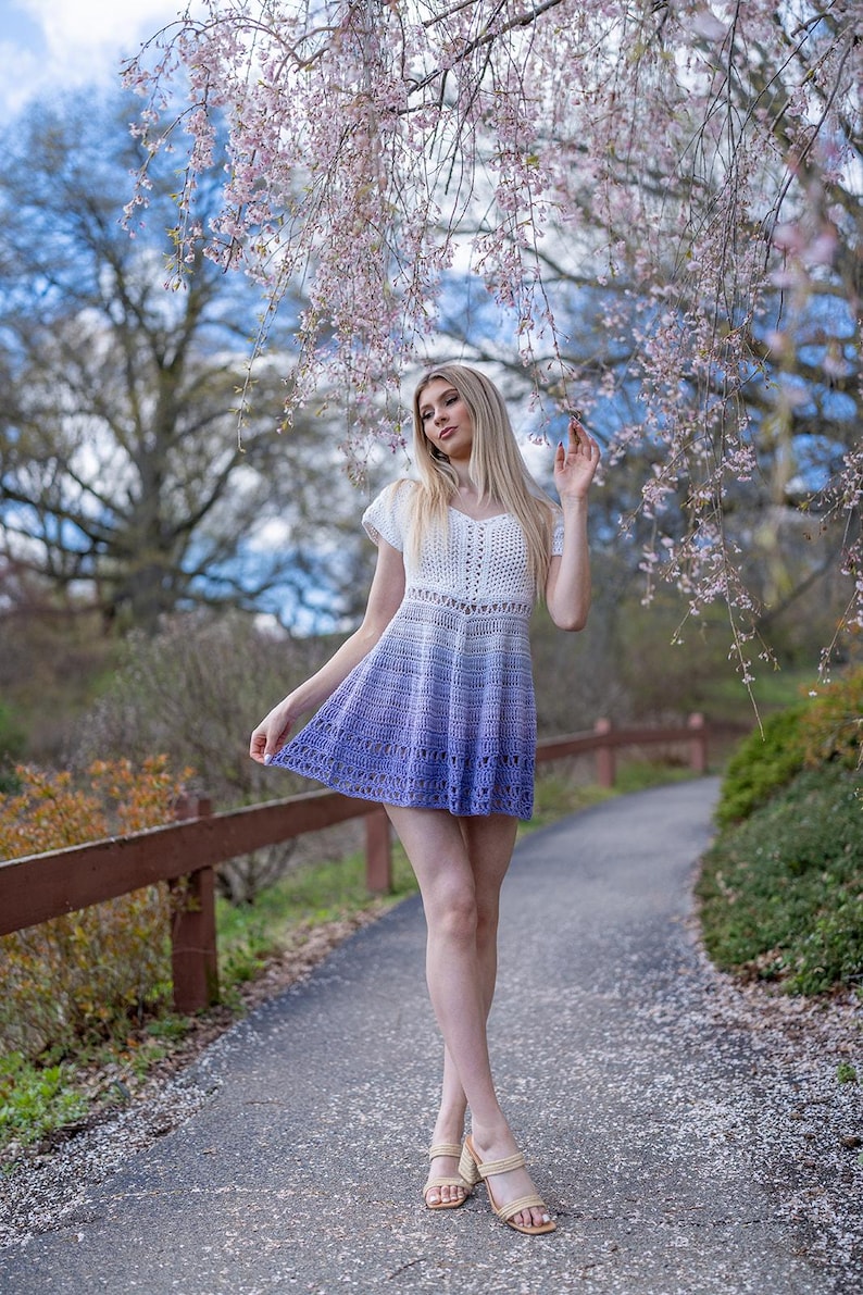 May include: A white and purple ombre crochet dress with short sleeves. The dress has a V-neck and a flared skirt. The model is wearing beige sandals. The background includes a path, a wooden fence, and flowering trees.