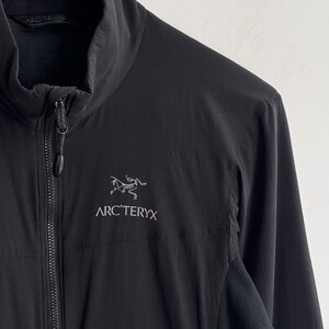 May include: Black jacket with a high collar and a full-length zipper. The jacket features the white Arc'teryx logo on the chest. The jacket has a textured panel on the side. The jacket is hanging on a black hanger.