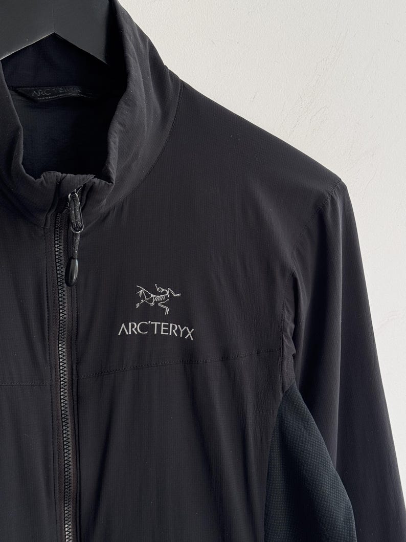 May include: Black jacket with a high collar and a full-length zipper. The jacket features the white Arc'teryx logo on the chest. The jacket has a textured panel on the side. The jacket is hanging on a black hanger.