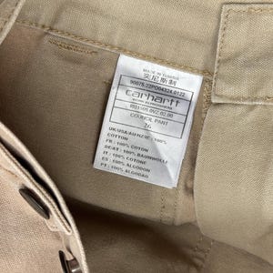 Puede incluir: Primer plano de pantalones Carhartt Council de color beige. La etiqueta dice "MADE IN TUNISIA" e incluye el logotipo de Carhartt e informaci&oacute;n del producto. Los pantalones son 100% algod&oacute;n. Talla 26.