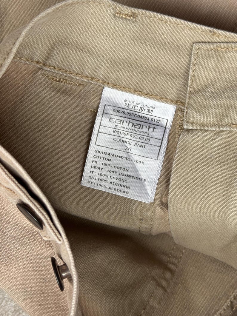 Puede incluir: Primer plano de pantalones Carhartt Council de color beige. La etiqueta dice "MADE IN TUNISIA" e incluye el logotipo de Carhartt e informaci&oacute;n del producto. Los pantalones son 100% algod&oacute;n. Talla 26.