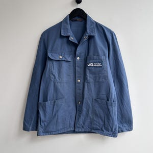 Può includere: Una giacca da lavoro blu con una toppa sul petto sinistro che recita "Kindt Wetzikon".