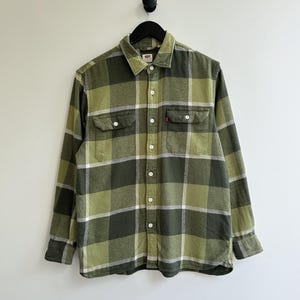Puede incluir: Camisa de franela a cuadros verdes y blancos con dos bolsillos en el pecho y cuello abotonado. La camisa tiene un corte clásico y una etiqueta roja de Levi's en el bolsillo. Cuelga de una percha negra contra una pared blanca.