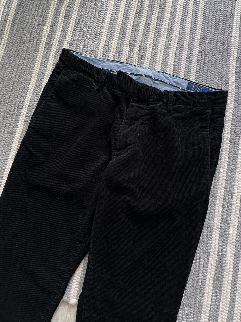 Puede incluir: Pantal&oacute;n de pana negro con una etiqueta amarilla y azul que dice "POLO RALPH LAUREN".