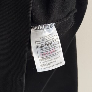May include: Close-up of a black Carhartt sweatshirt with a white tag. The tag displays the brand name, product information, and the text "SHATTER SWEAT". The fabric composition is listed as 58% cotton and 42% polyester.