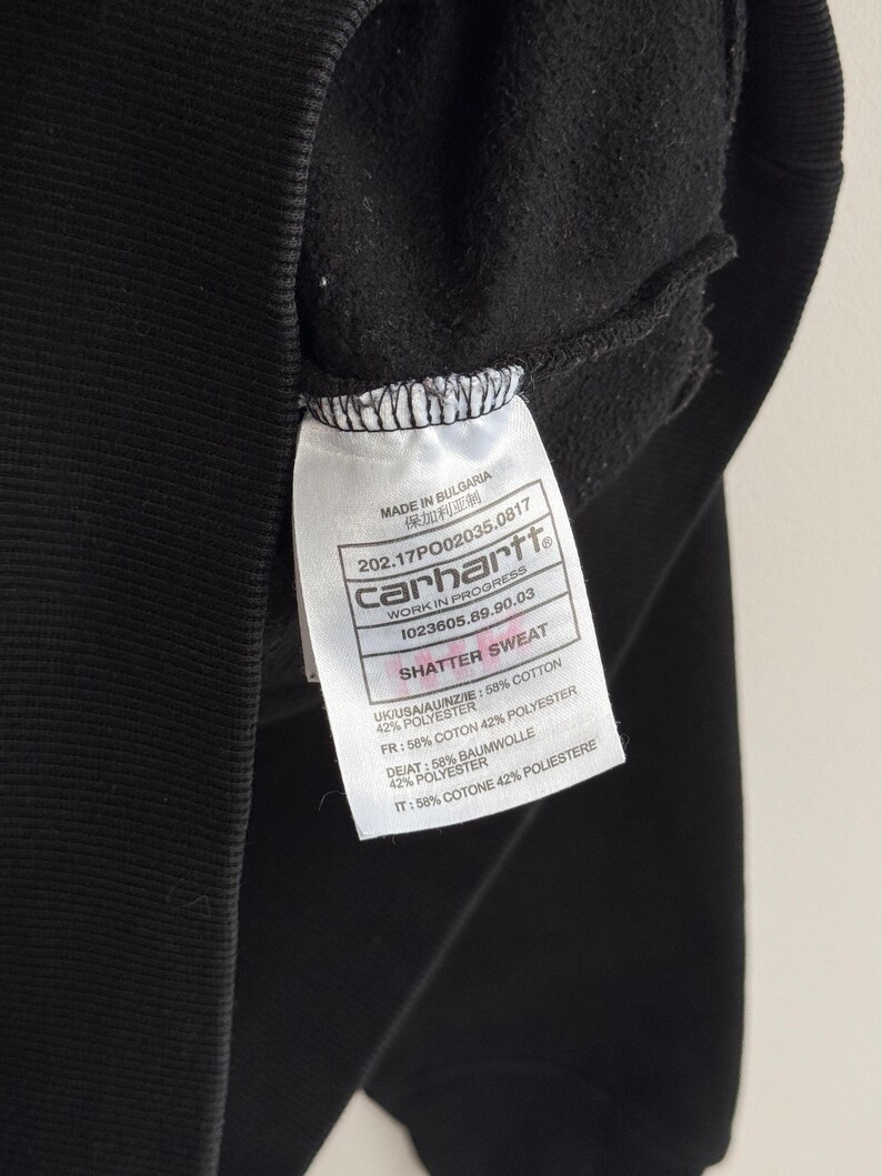 May include: Close-up of a black Carhartt sweatshirt with a white tag. The tag displays the brand name, product information, and the text "SHATTER SWEAT". The fabric composition is listed as 58% cotton and 42% polyester.