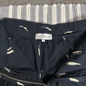 Puede incluir: Primer plano de unos pantalones cortos negros con patrones blancos en forma de pinceladas. Los pantalones cortos tienen cierre de cremallera y una etiqueta que dice "Gianni Versace MADE IN ITALY". La prenda es talla II.