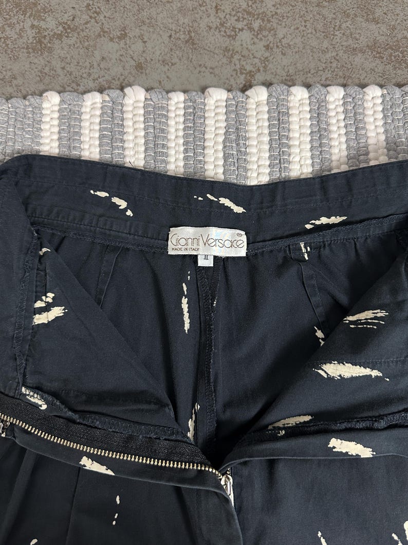 Puede incluir: Primer plano de unos pantalones cortos negros con patrones blancos en forma de pinceladas. Los pantalones cortos tienen cierre de cremallera y una etiqueta que dice "Gianni Versace MADE IN ITALY". La prenda es talla II.