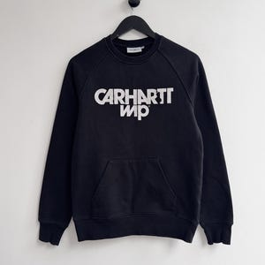 May include: Black sweatshirt with the white text "CARHARTT WIP" across the chest. The sweatshirt has a kangaroo pocket and raglan sleeves. The garment is hanging on a black hanger against a white background.