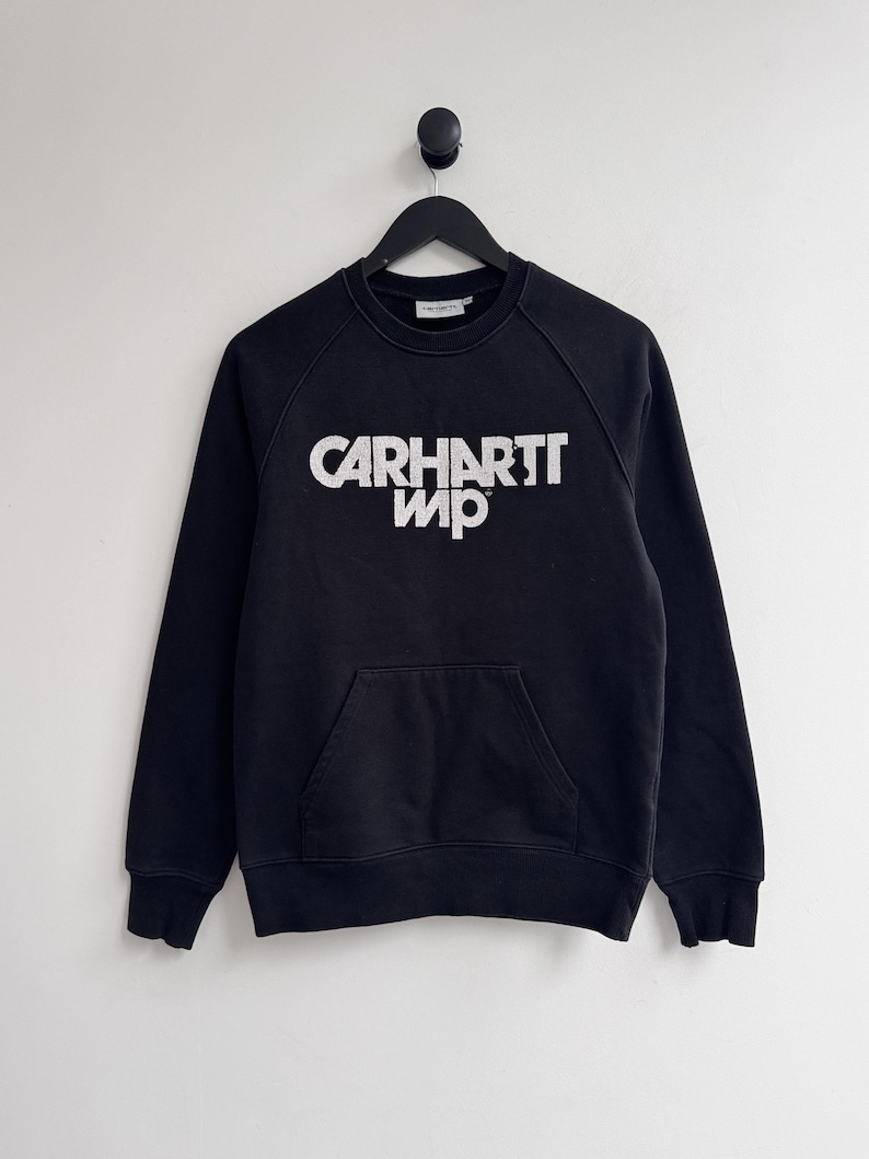May include: Black sweatshirt with the white text "CARHARTT WIP" across the chest. The sweatshirt has a kangaroo pocket and raglan sleeves. The garment is hanging on a black hanger against a white background.