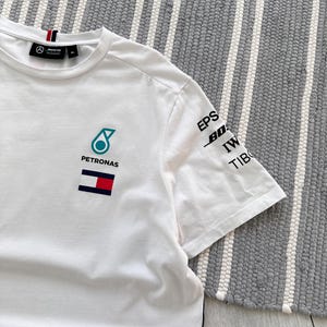 May include: White t-shirt with a blue and green Petronas logo, a Tommy Hilfiger logo, and black text on the right sleeve that says "EPSON, BOSCH, IWC, TIBCO".