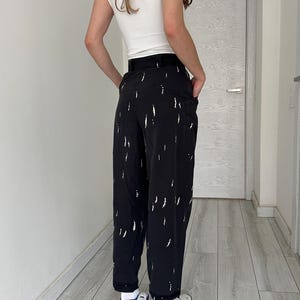 Puede incluir: Pantalones negros con dise&ntilde;o de pinceladas blancas. Los pantalones tienen un corte holgado y cintura alta. La persona lleva una camiseta blanca y zapatillas blancas y azules. Los pantalones tienen bolsillos.