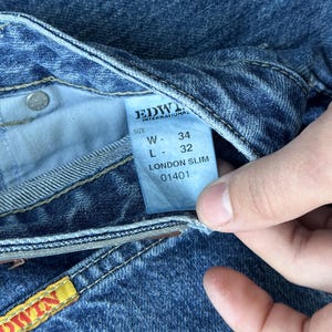 Pode incluir: Close-up de jeans azuis com uma etiqueta que diz "EDWIN INTERNATIONAL" e "LONDON SLIM". A etiqueta tamb&eacute;m indica uma cintura de 86 cm e um comprimento de 81 cm. Os jeans t&ecirc;m um design cl&aacute;ssico de cinco bolsos.