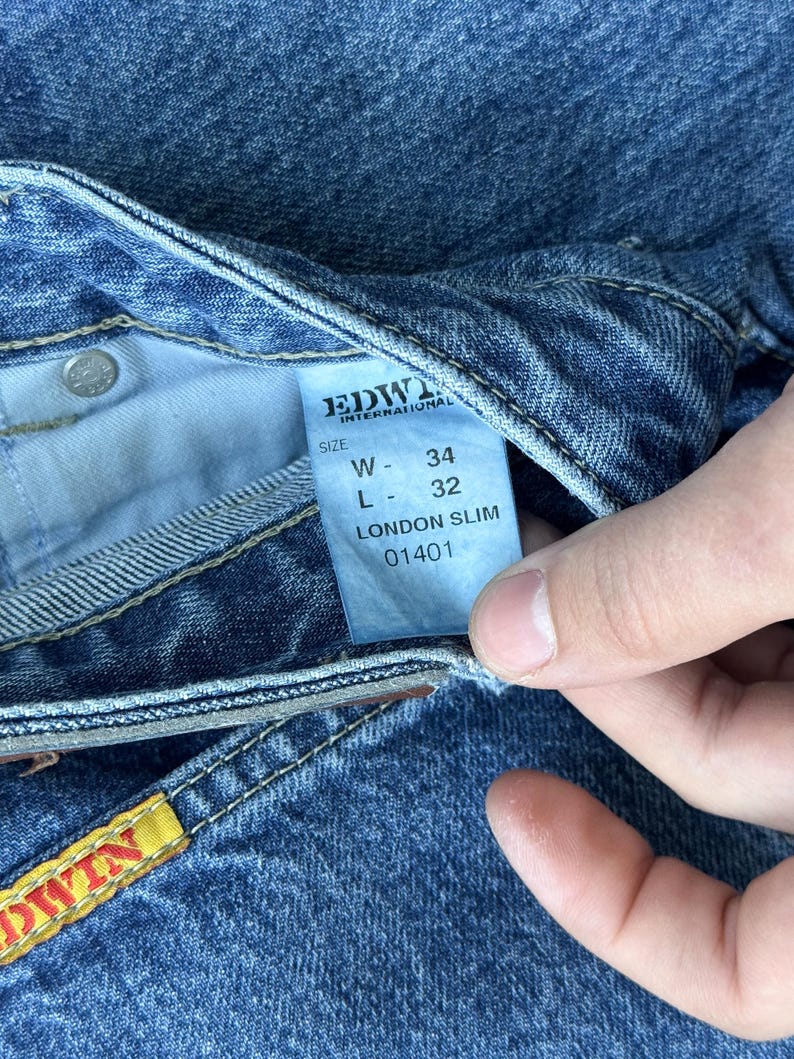 Pode incluir: Close-up de jeans azuis com uma etiqueta que diz "EDWIN INTERNATIONAL" e "LONDON SLIM". A etiqueta tamb&eacute;m indica uma cintura de 86 cm e um comprimento de 81 cm. Os jeans t&ecirc;m um design cl&aacute;ssico de cinco bolsos.