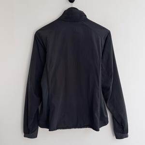 May include: Black jacket with a high collar and long sleeves. The jacket is hanging on a black hook against a white wall. The jacket has a full zipper and a slightly curved hemline.