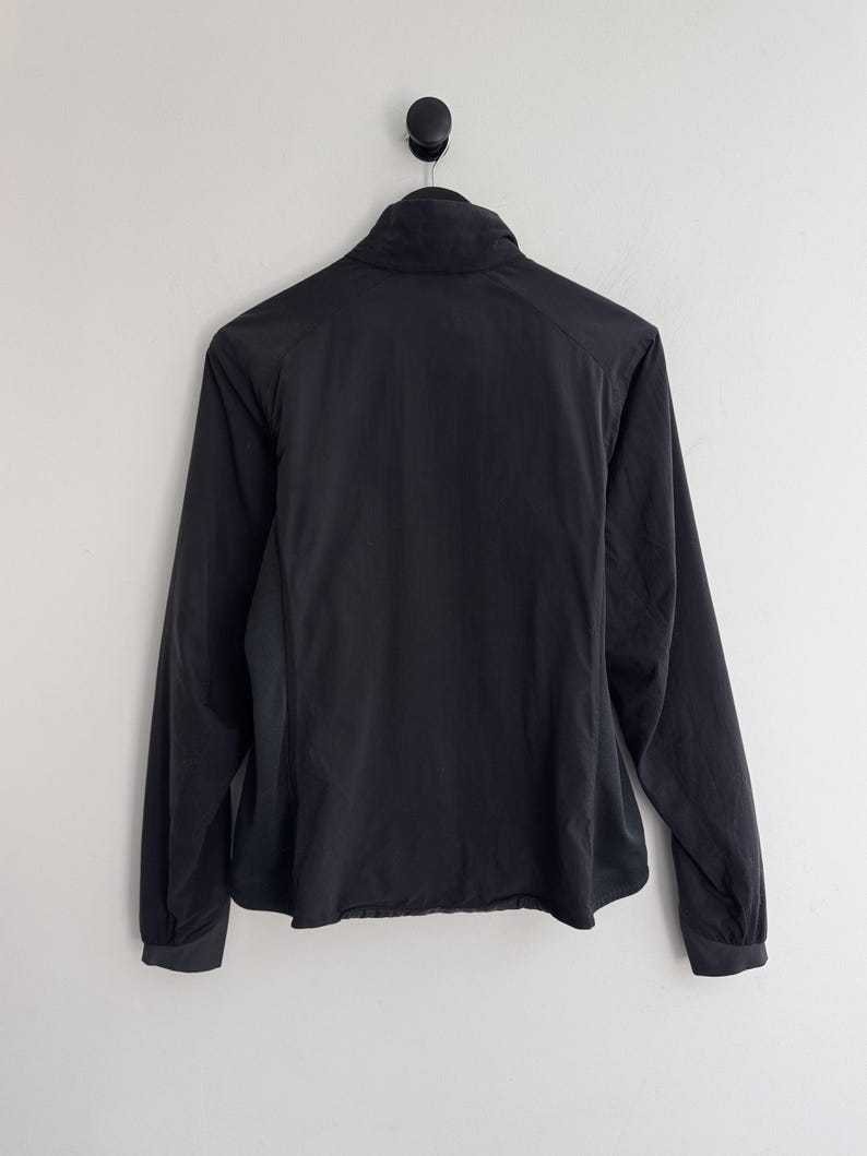 May include: Black jacket with a high collar and long sleeves. The jacket is hanging on a black hook against a white wall. The jacket has a full zipper and a slightly curved hemline.