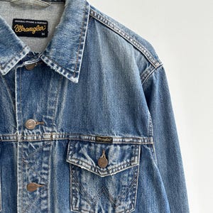 May include: A blue denim jacket with a faded look. The jacket has a button closure, two front pockets, and a chest pocket. The label on the jacket reads "Wrangler" with the text "Original Styling & Features" above it.
