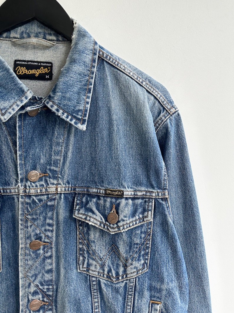 May include: A blue denim jacket with a faded look. The jacket has a button closure, two front pockets, and a chest pocket. The label on the jacket reads "Wrangler" with the text "Original Styling & Features" above it.