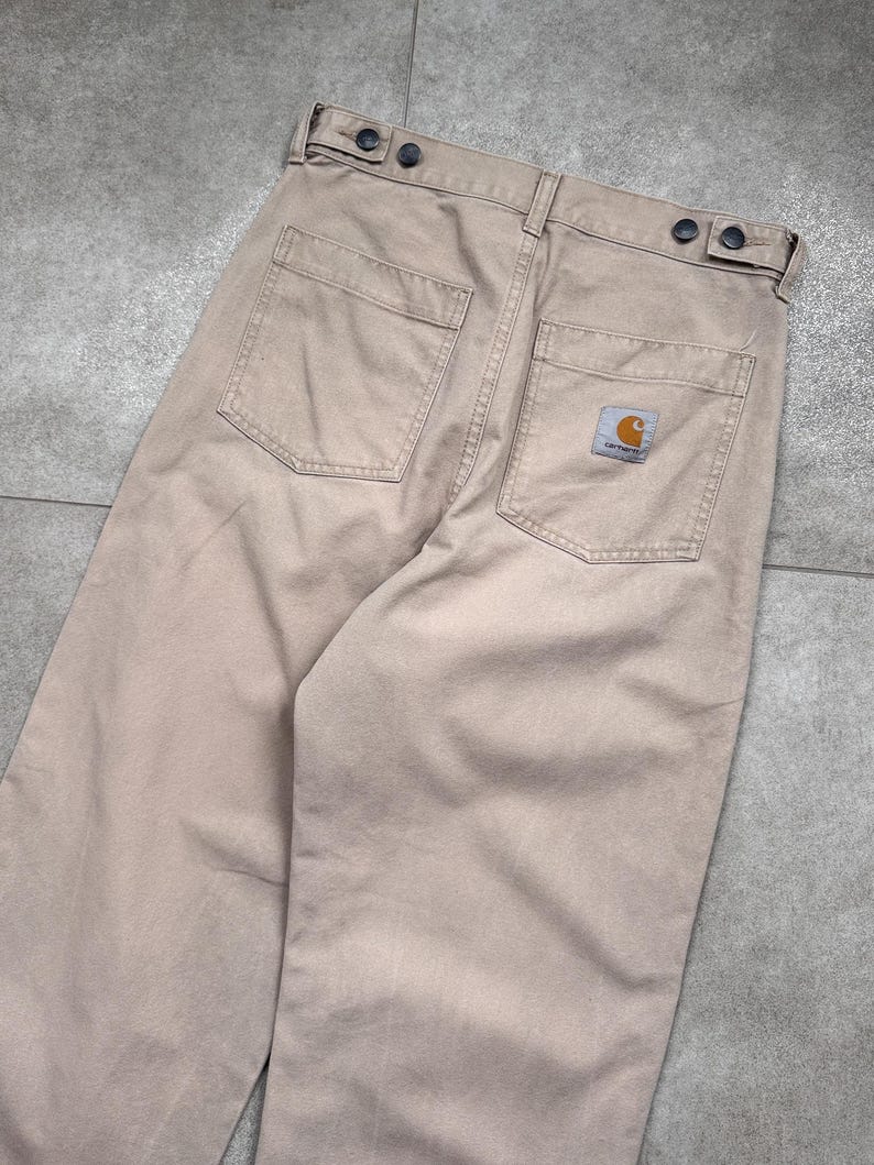 Puede incluir: Pantalones de trabajo color beige con dos bolsillos traseros y un peque&ntilde;o parche con el logotipo. Los pantalones tienen cierre de bot&oacute;n y un dise&ntilde;o de pierna recta. La tela parece duradera y adecuada para el trabajo o el uso casual.